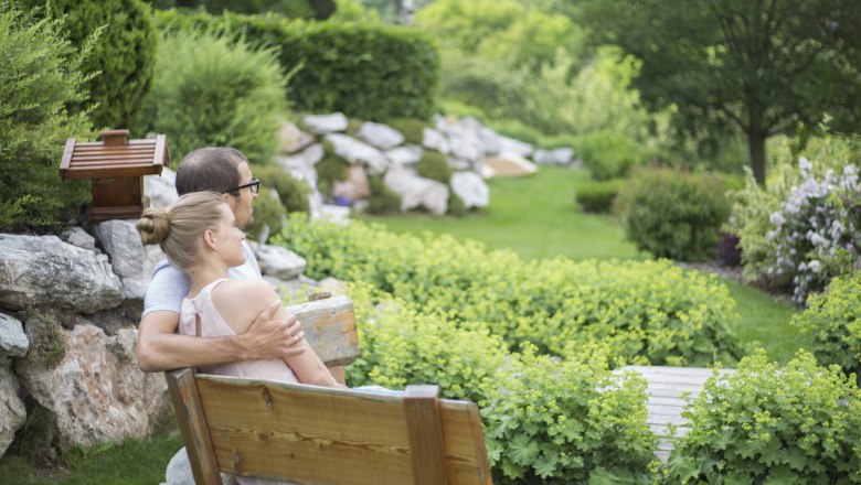 Enjoy the peace and quiet in the alpine garden., © Patrick Haberler A couple is sitting on a wooden bench in a green garden with bushes and trees.