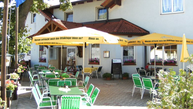 Gastgarten, © Martin Zwinz Ein Gastgarten mit grünen Stühlen und gelben Sonnenschirmen vor einem Gebäude.