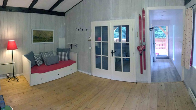 Ferienhaus, © Marianne Gschaider Innenansicht eines gemütlichen Ferienhauses mit Holzboden, rotem Sofa, Skiern an der Wand und Glastüren.