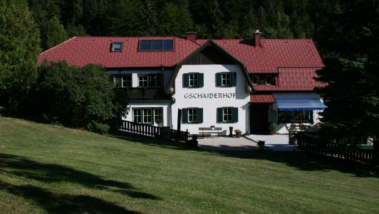Frühstückspension Gschaiderhof, © Marianne Gschaider Ein traditionelles Gebäude mit rotem Dach und grünen Fensterläden, umgeben von Bäumen und Wiese, mit dem Schriftzug 'Gschaiderhof'.