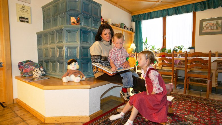 Entspannen am Kamin, © Hausmann/Zwickl Eine Frau sitzt mit zwei Kindern auf einer Bank vor einem Kachelofen und liest ein Buch. Spielzeug und Dekorationen sind um sie herum verteilt.
