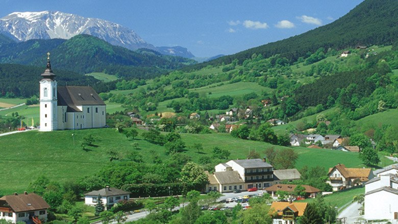 Der Landgasthof Jautschnig an der Wallfahrtskirche Maria Kirchbüchl, © Gasthaus Jautschnig Landschaft mit Kirche auf Hügel, umgeben von grünen Feldern und Bergen im Hintergrund.
