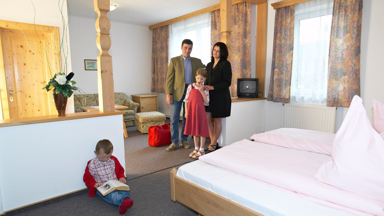 Die gemütlichen Zimmer, © Hausmann/Zwickl Familie in einem gemütlichen Hotelzimmer mit Holzmöbeln und rosa Bettwäsche.