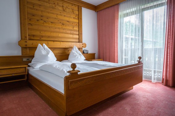 Gemütlich schlafen im Bruckerhof, © Wiener Alpen / Christian Kremsl Ein gemütliches Schlafzimmer mit einem Doppelbett aus Holz, weißen Bettdecken und roten Vorhängen vor einem Fenster.