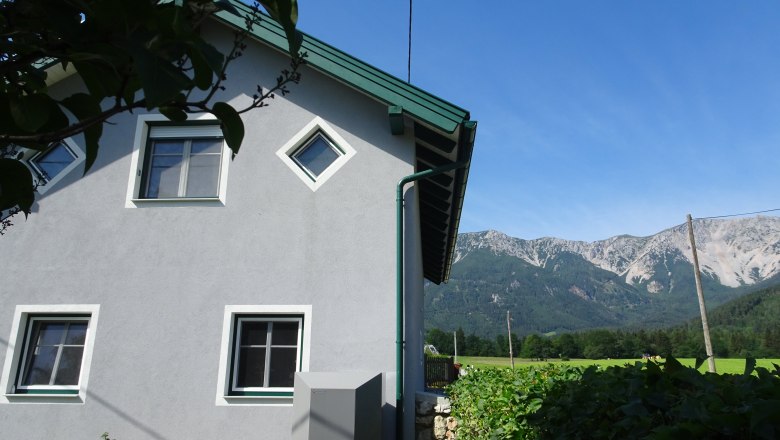 Ferienwohnung Schneebergblick, © Familie Zwinz Haus mit grauer Fassade und grünen Fensterrahmen, im Hintergrund Berge und blauer Himmel.