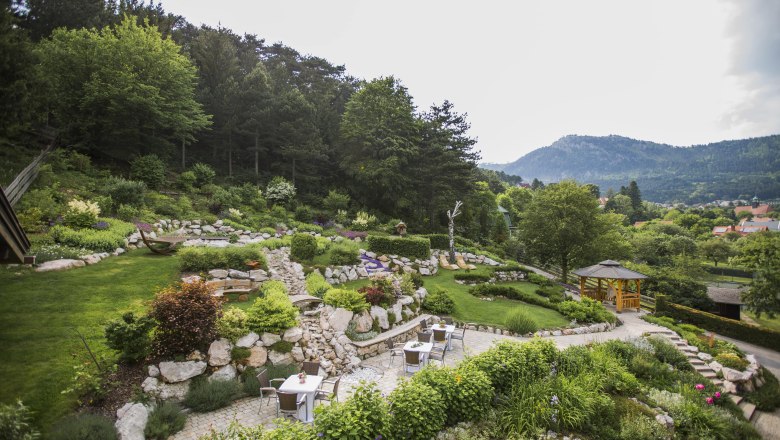 Alpengarten, © Patrick Haberler Ein terrassierter Garten mit Steinen, Pflanzen und Sitzbereichen, umgeben von Bäumen und Bergen im Hintergrund.