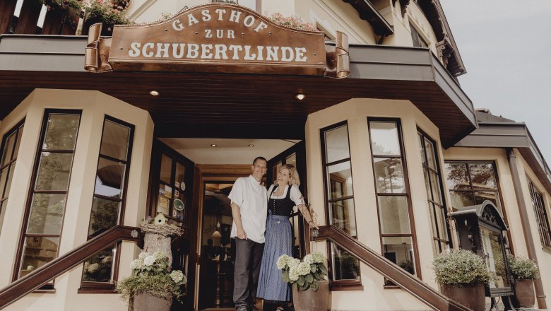 „Zur Schubertlinde“ im Schneebergland, © Niederösterreich Werbung/Rita Newman Eingang des Gasthofs Zur Schubertlinde mit zwei Personen in traditioneller Kleidung.