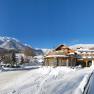 Winterlandschaft mit Hotel Forellenhof vor schneebedeckten Bergen und blauem Himmel.