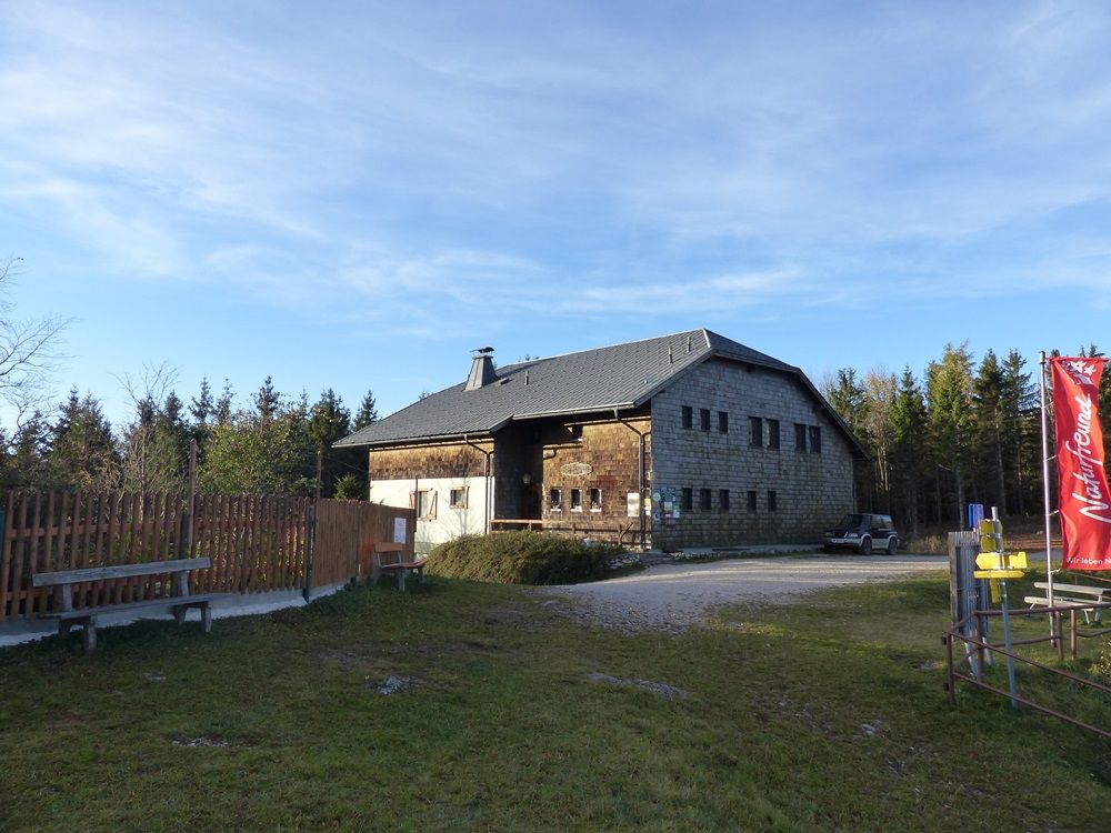 Ein rustikales Gebäude mit Holzwänden und einem grauen Dach, umgeben von Bäumen. Eine rote Fahne steht rechts im Bild.