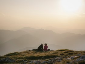 Zwei Personen sitzen im Abendlicht auf einer Wiese am Schneeberg und blicken &uuml;ber die Berglandschaft