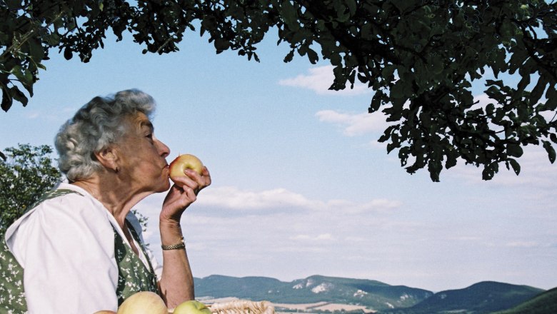Ältere Frau mit Korb voller Äpfel, die einen Apfel küsst, vor einer Landschaft mit Hügeln und blauem Himmel.
