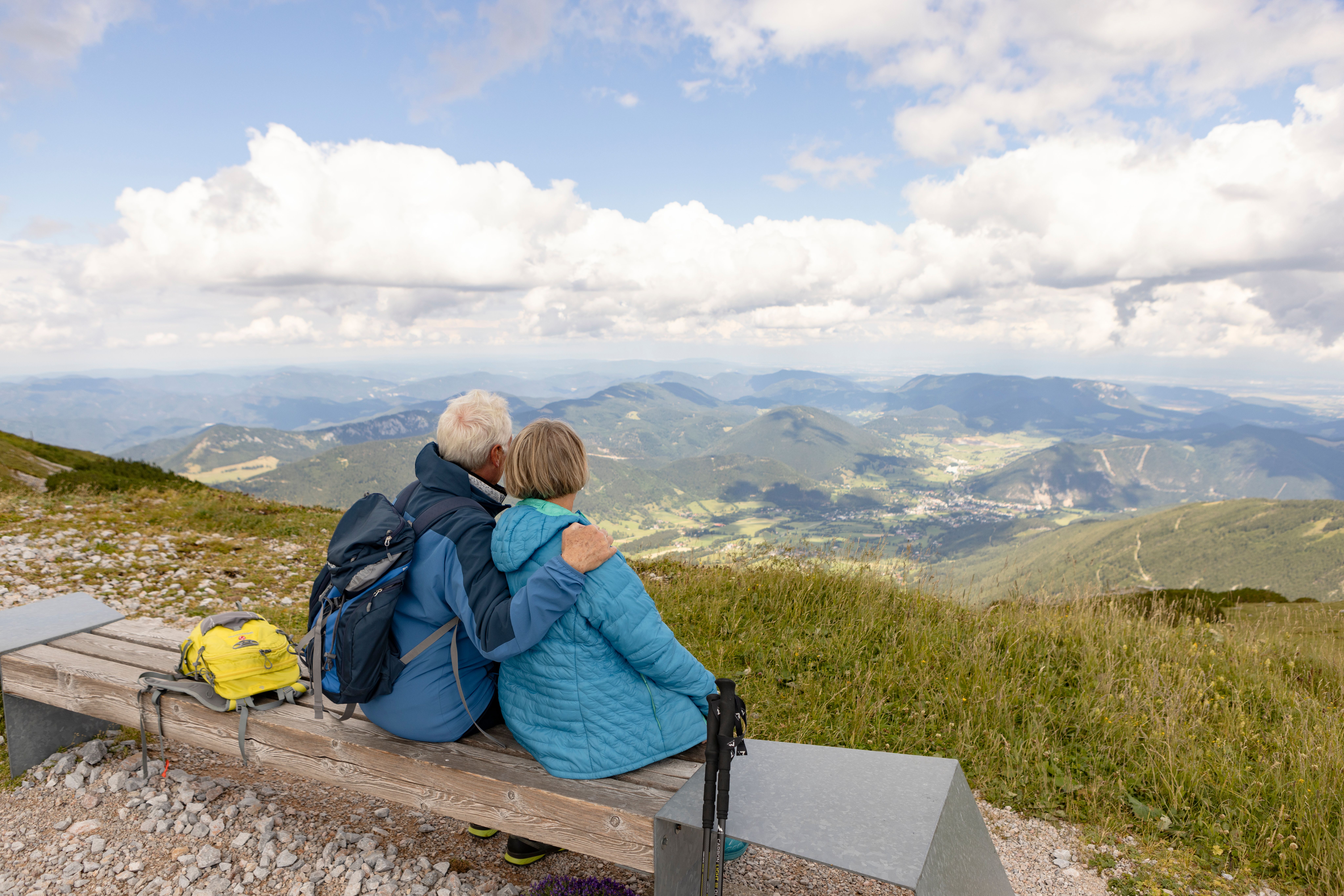 Älteres Paar in Outdoorjacken sitzt auf einer Bank auf einem Berg und blickt auf ein weites Tal mit Bergen unter bewölktem Himmel