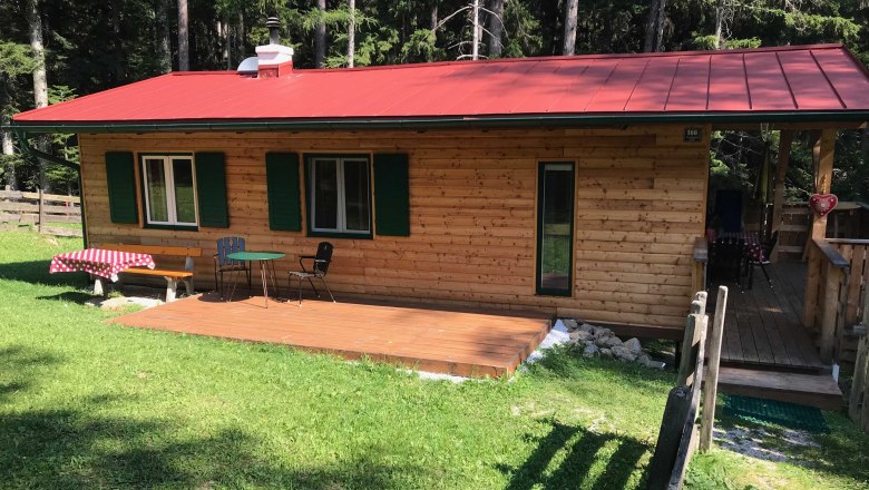 A small wooden holiday home with a red roof, terrace and garden furniture, surrounded by forest.