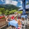 Familie auf sonniger Terrasse mit Bergen im Hintergrund, bedient von Kellnerin in traditioneller Kleidung.