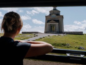 Eine Frau blickt von der Schneebergbahn auf das Elisabethkircherl am Hochschneeberg. Ein Kiesweg f&uuml;hrt zur Kirche, die umgeben von einer gr&uuml;nen Wiese ist, was eine Atmosph&auml;re der Erkundung und historischer Neugier erzeugt.
