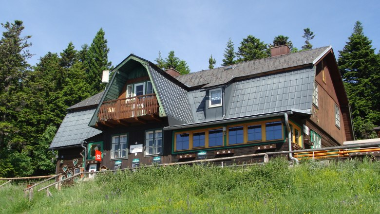A traditional mountain house with a wooden fa&ccedil;ade and shingle roof, surrounded by trees and meadows.