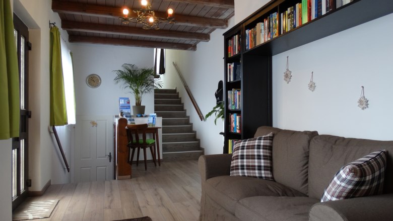 Wohnzimmer mit braunem Sofa, B&uuml;cherregal und Treppe im Hintergrund.