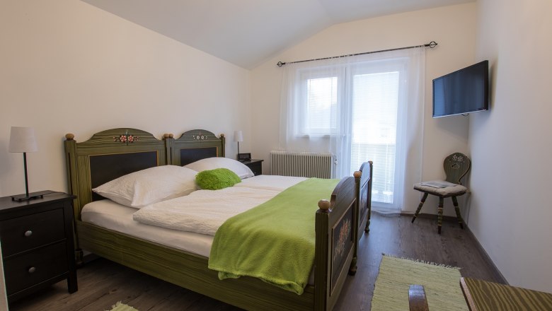 Gem&uuml;tliches Schlafzimmer mit Doppelbett, gr&uuml;ner Bettdecke und TV an der Wand.