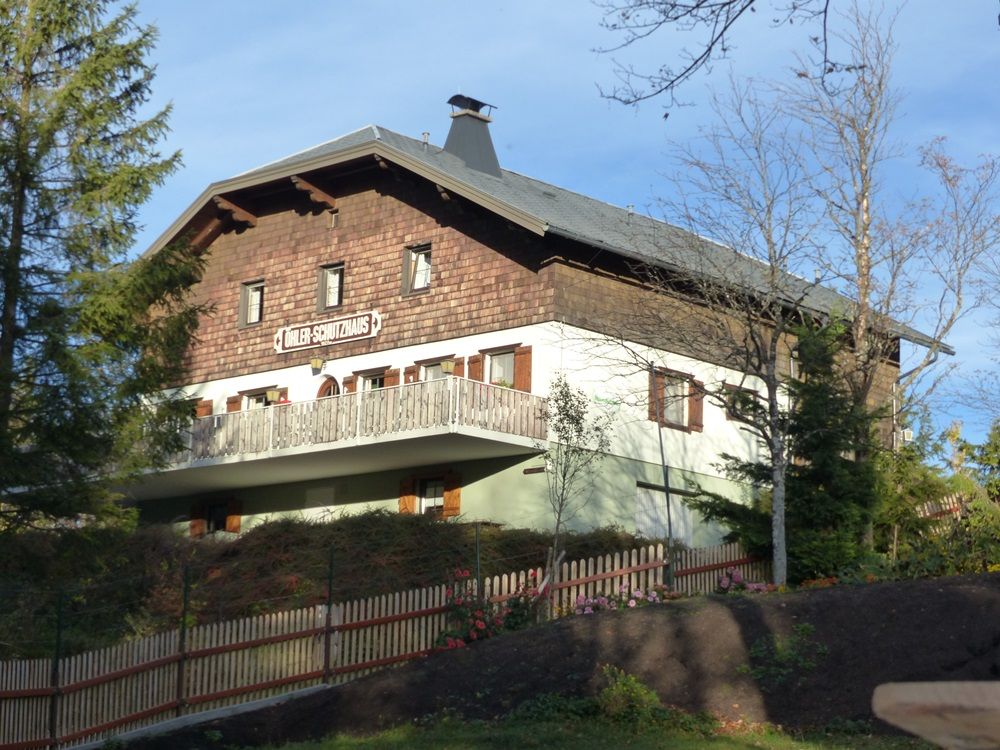 Ein traditionelles Gebäude mit Holzverkleidung und Balkon, umgeben von Bäumen und einem Holzzaun. Schild: "Döhler-Schutzhaus".
