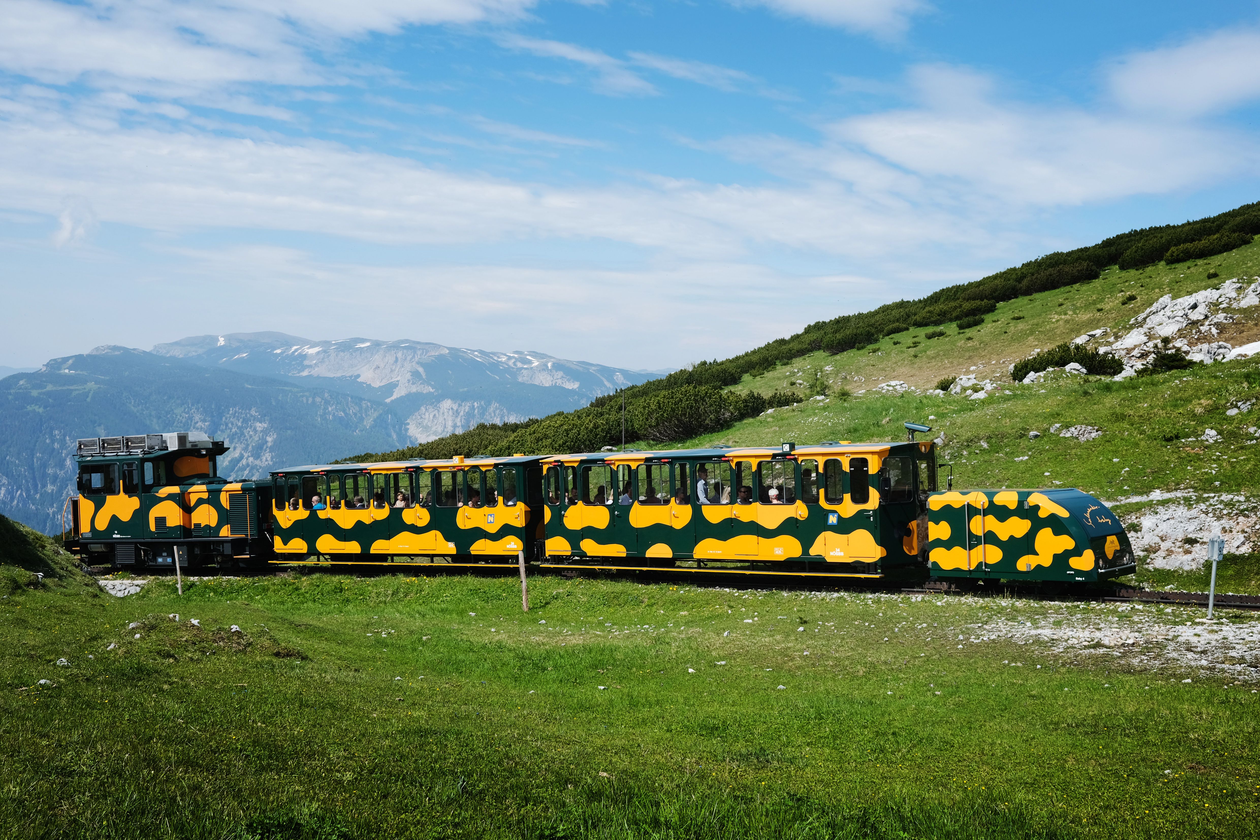 Gelb-grüner Zug fährt über einen Berghang. Dahinter erstreckt sich eine alpine Landschaft mit grünen Wiesen, Felsen und weitläufigem Bergpanorama.