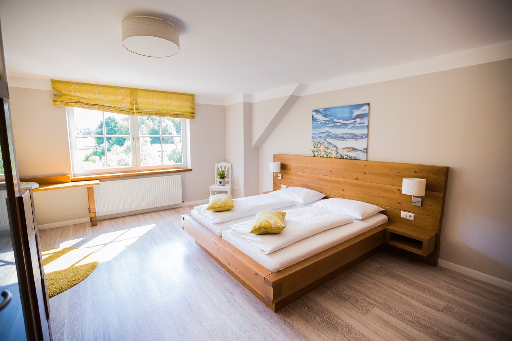 Helles Doppelzimmer mit Holzbett, gelben Kissen und großem Fenster.