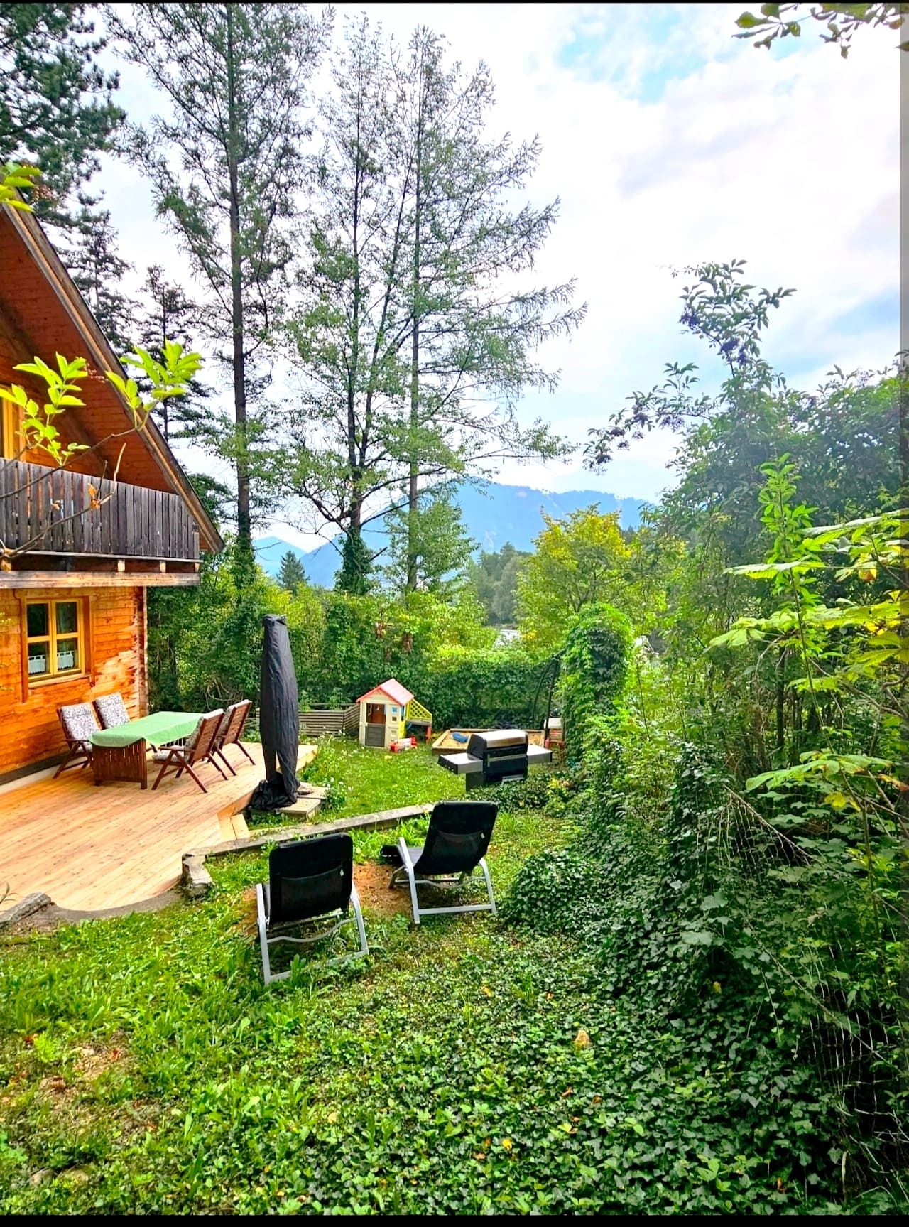 Ein Ferienhaus mit Terrasse und Gartenmöbeln, umgeben von Bäumen und grüner Vegetation. Im Hintergrund sind Berge zu sehen.