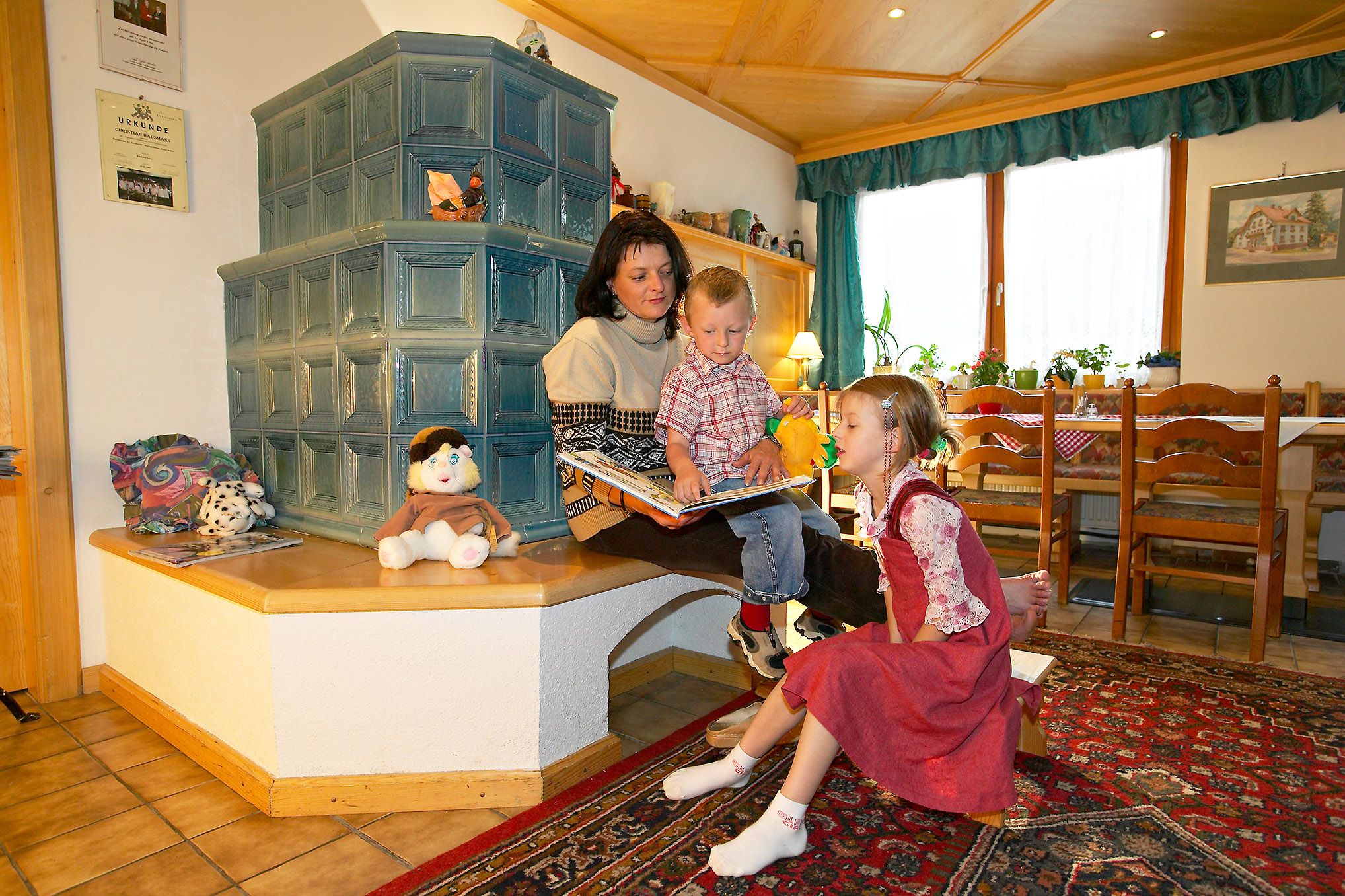Eine Frau sitzt mit zwei Kindern auf einer Bank vor einem Kachelofen und liest ein Buch. Spielzeug und Dekorationen sind um sie herum verteilt.