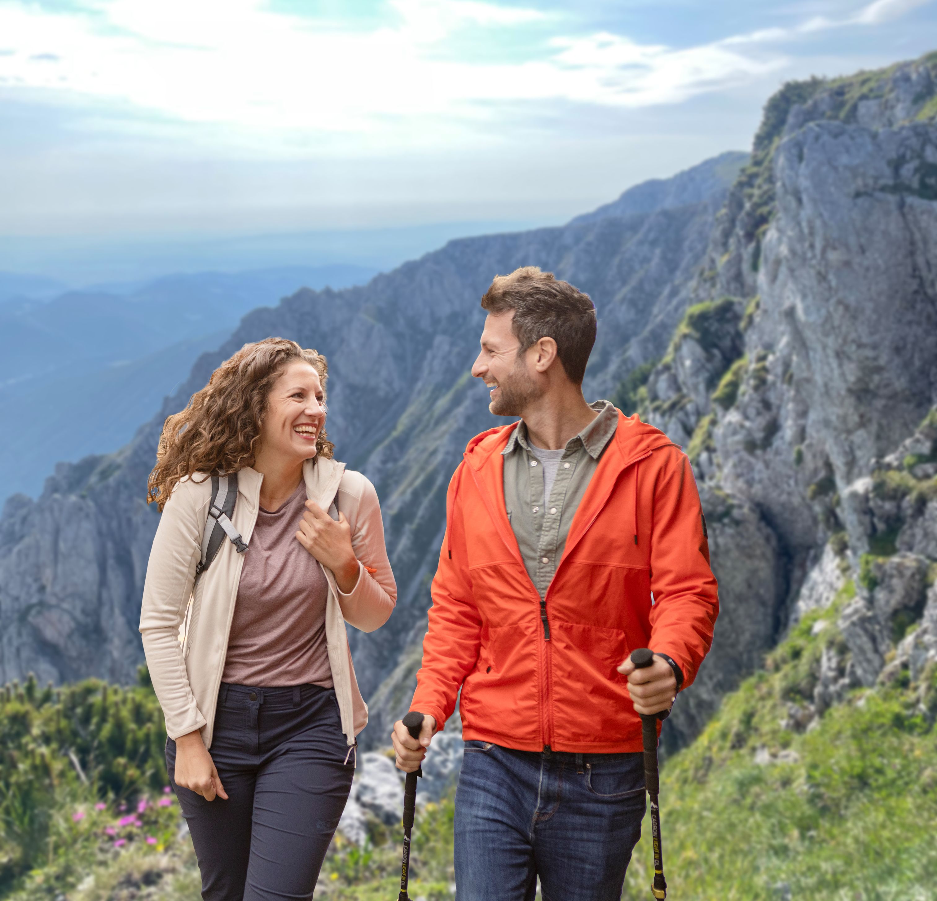 Ein lächelndes Paar wandert bei strahlendem Sonnenschein in den Bergen, schaut sich gut gelaunt an und trägt Outdoor-Kleidung sowie Wanderstöcke vor einer imposanten Felskulisse.