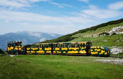 Gelb-grüner Zug fährt über einen Berghang. Dahinter erstreckt sich eine alpine Landschaft mit grünen Wiesen, Felsen und weitläufigem Bergpanorama.