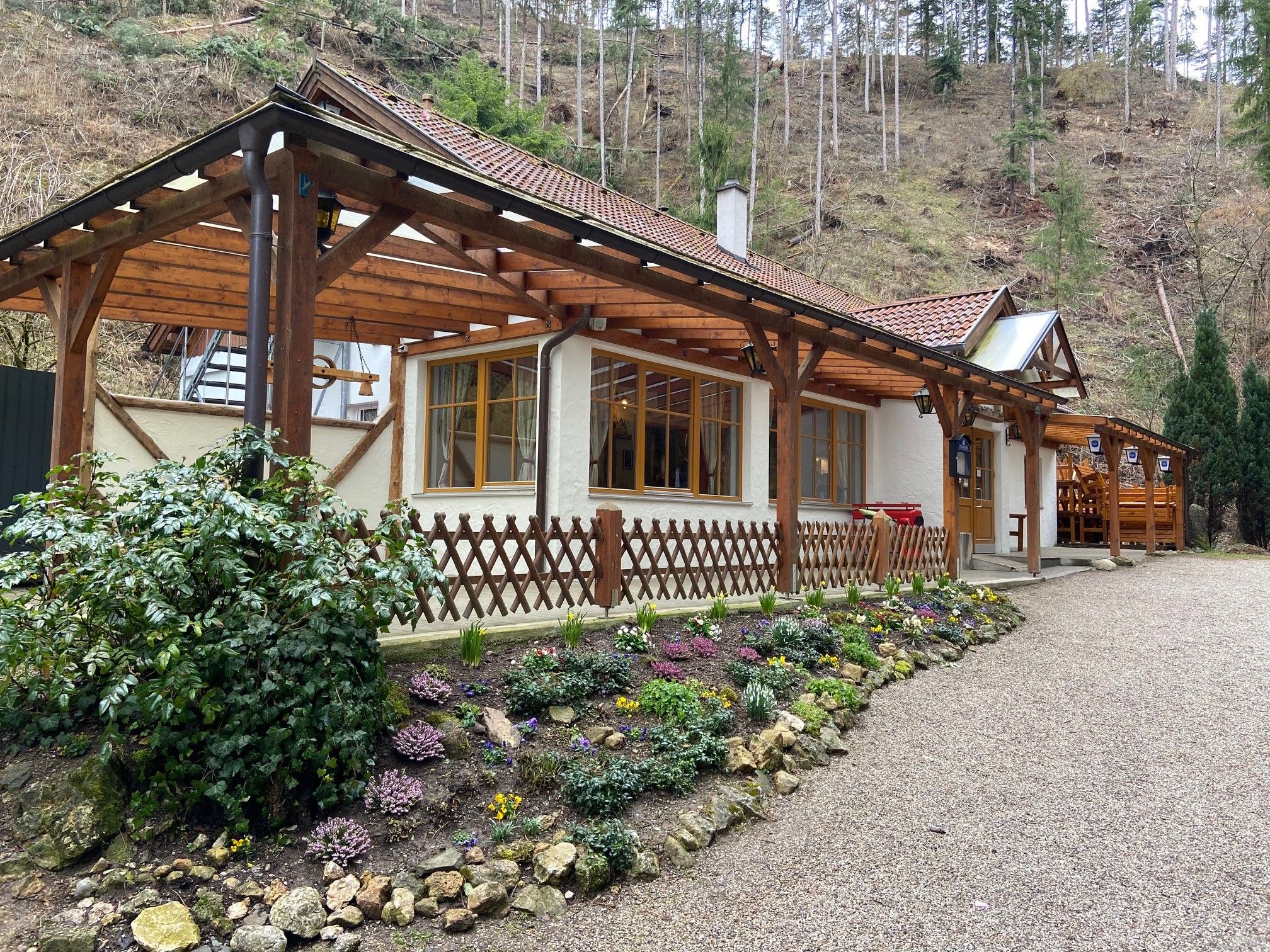 Ein Gasthaus mit Holzdach und Blumengarten vor einem bewaldeten Hang.