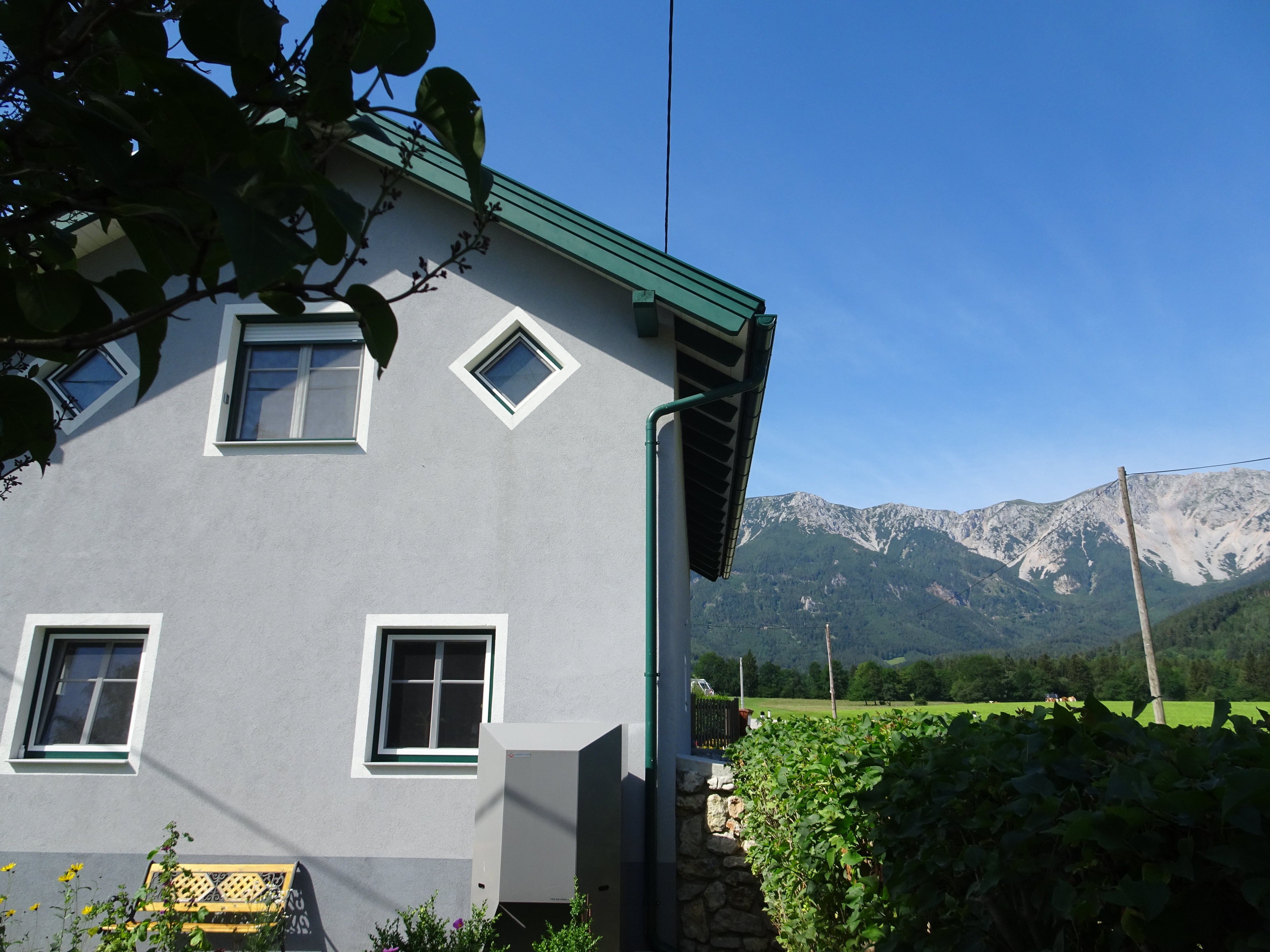 Haus mit grauer Fassade und grünen Fensterrahmen, im Hintergrund Berge und blauer Himmel.