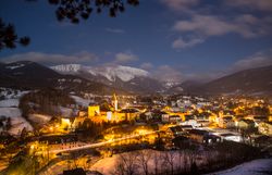 Winterliche Nachtaufnahme einer beleuchteten Bergstadt mit schneebedeckten Hügeln und Bergen im Hintergrund