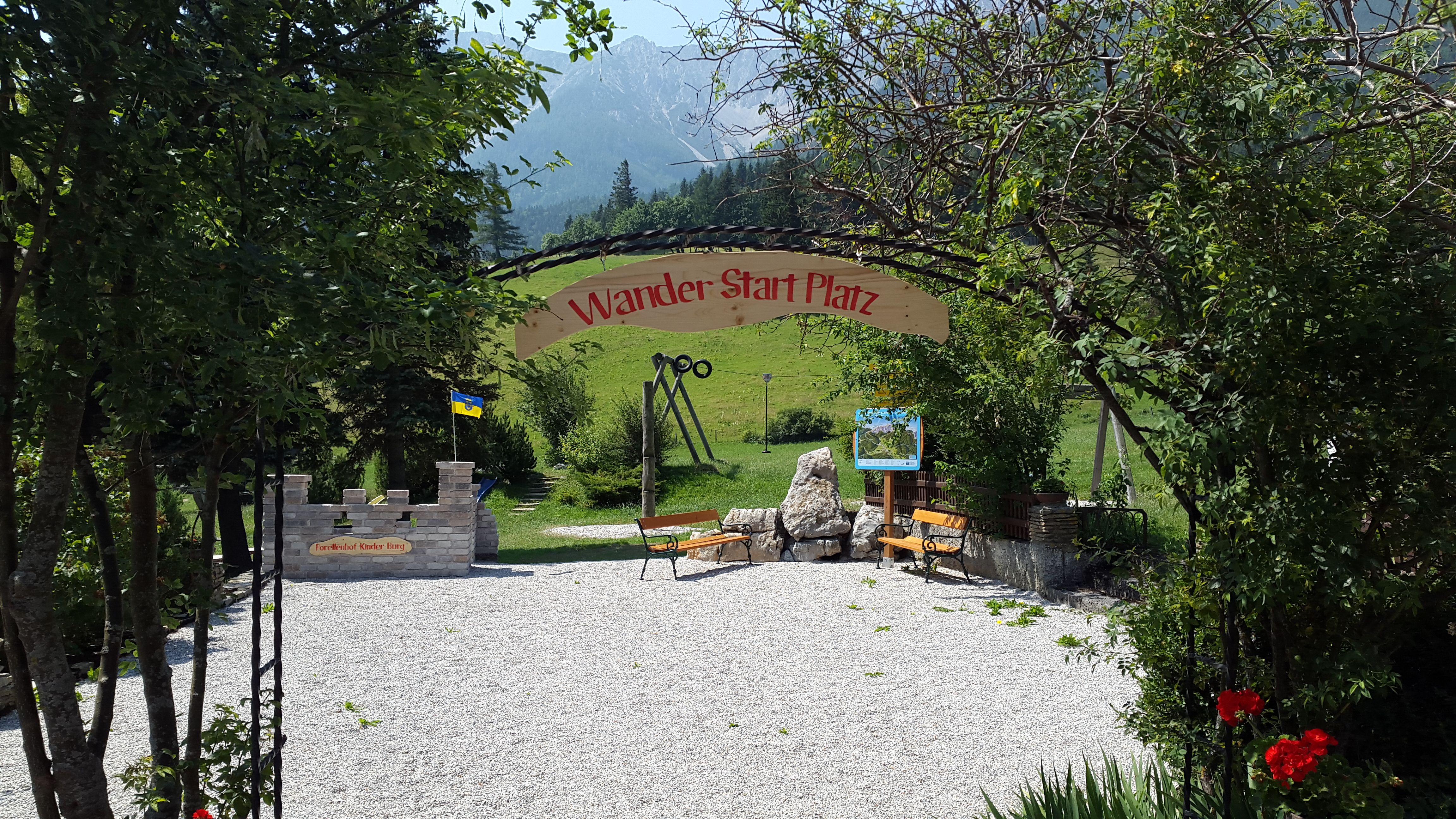 Eingang zu einem Wanderstartplatz mit Bänken und einem Schild, umgeben von Bäumen und Bergen im Hintergrund.