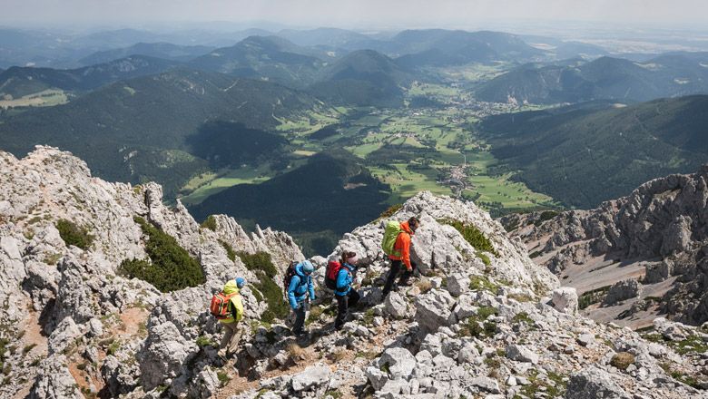 Gruppe von Wanderern auf einem felsigen Bergpfad mit Blick auf eine grüne Talebene und umliegende Berge.