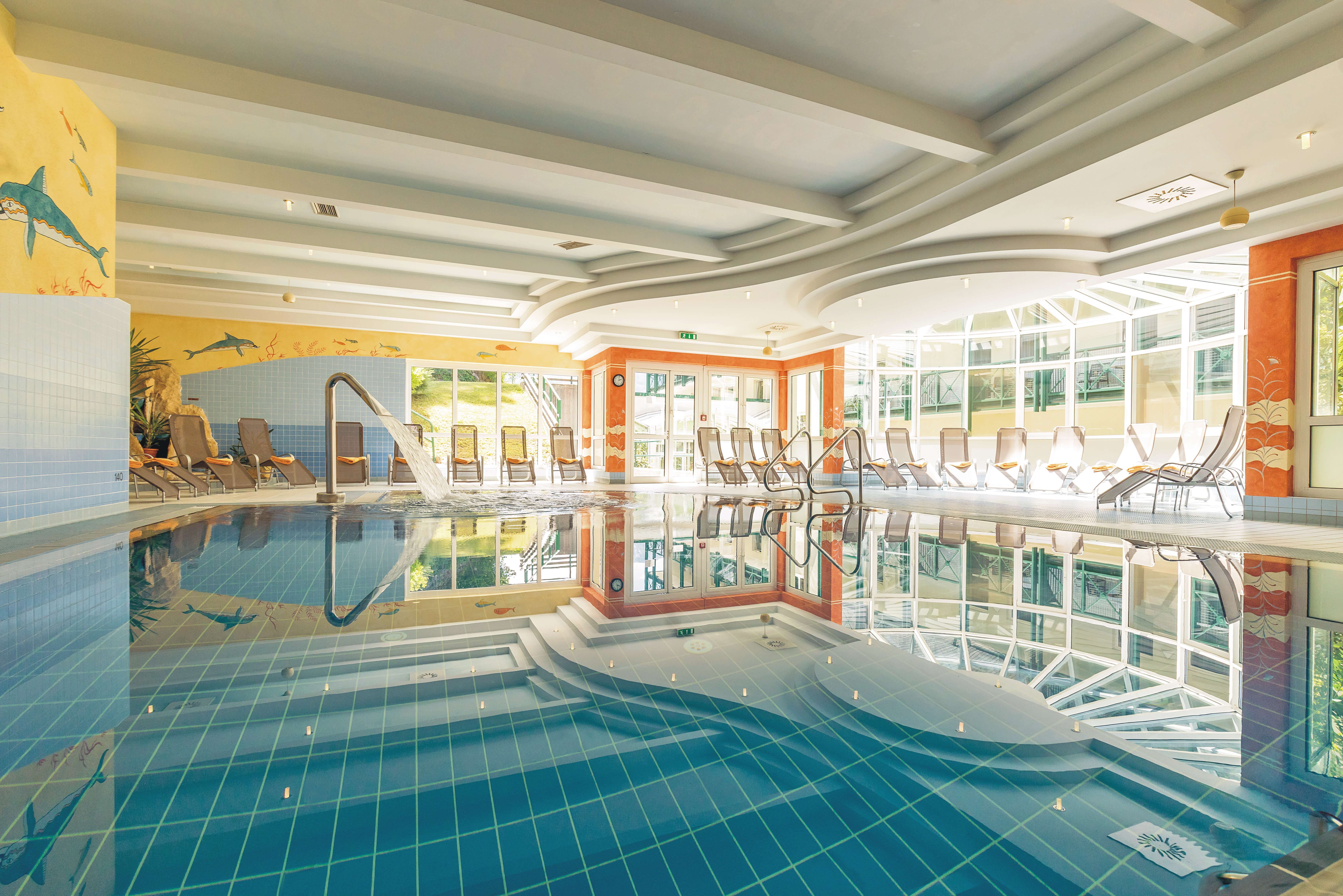Ein helles Hallenbad mit Liegestühlen und einem Wasserstrahl im Pool.