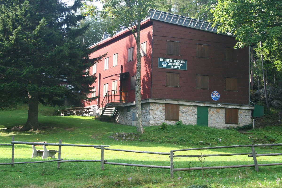 Ein rotes Gebäude mit Holzverkleidung, umgeben von Bäumen und einer Wiese, mit einem Schild "Naturfreundehaus Ortsgruppe Ternitz".