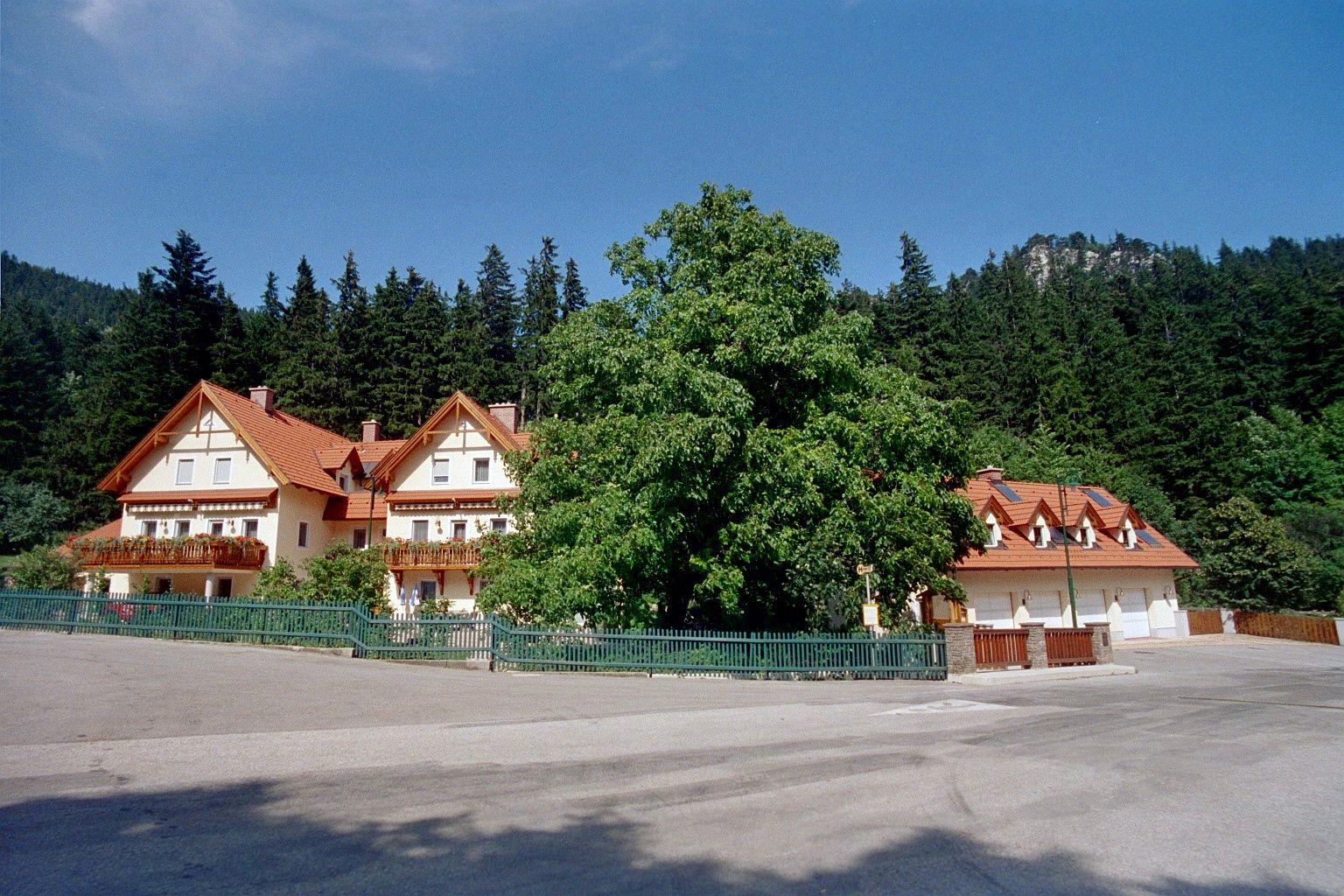 Ein großes Haus mit roten Dächern und Balkonen, umgeben von Bäumen und einem Wald im Hintergrund.