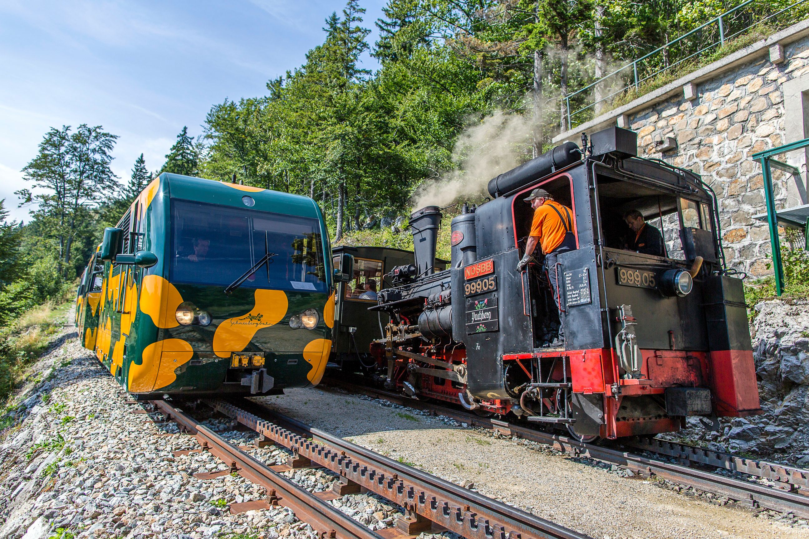 Moderner gelb-grüner Salamanderzug und historische Dampflokomotive begegnen sich auf einer Bergstrecke. Im Hintergrund stehen Wald und eine steinerne Stützmauer.