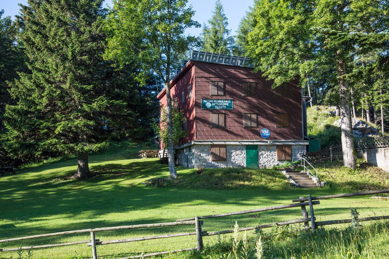 Eine Berghütte umgeben von Bäumen und Wiese, mit einem Holzzaun im Vordergrund.