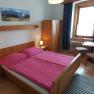 A cozy double room with wooden furniture, a double bed with pink bed linen, a table with two chairs and a small TV.