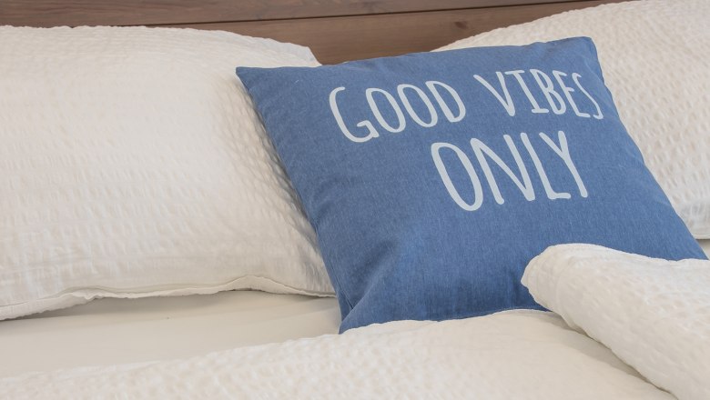 Bett mit blauem Kissen, Aufschrift 'Good Vibes Only'.