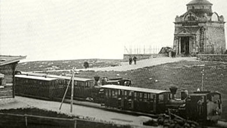 Schwarz-weiße Aufnahme einer historischen Bergbahn mit mehreren Waggons und einem Gebäude auf einem Berggipfel