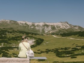 Frau sitzt auf einer Bank und genie&szlig;t den Ausblick auf die Berge beim Damb&ouml;ckhaus am Schneeberg