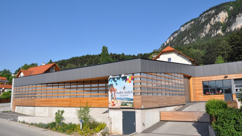 Modernes Gebäude mit Holzverkleidung und Werbeschild von Mohr-Sederl Fruchtwelt, umgeben von grüner Landschaft und Bergen.