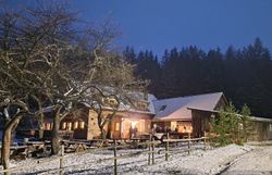 Winterliche Hofszene mit schneebedecktem Boden, beleuchtetem Bauernhaus und kahlen Bäumen vor dunklem Waldhintergrund