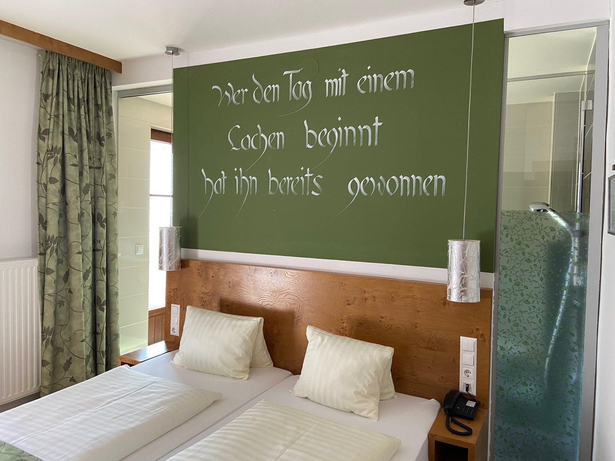 Ein Doppelzimmer mit zwei Betten, grüner Wand mit Spruch, Vorhängen und einer Dusche mit Glaswand.