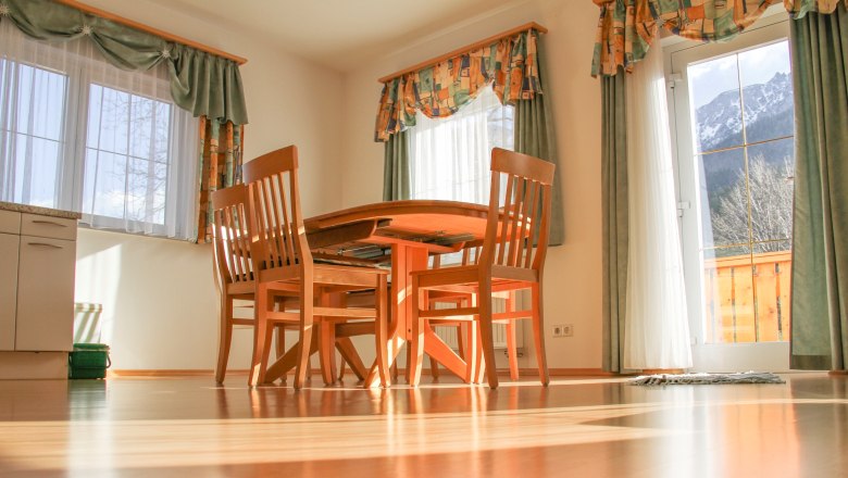Dining area with wooden table and chairs in a sunny room with large windows and curtains.