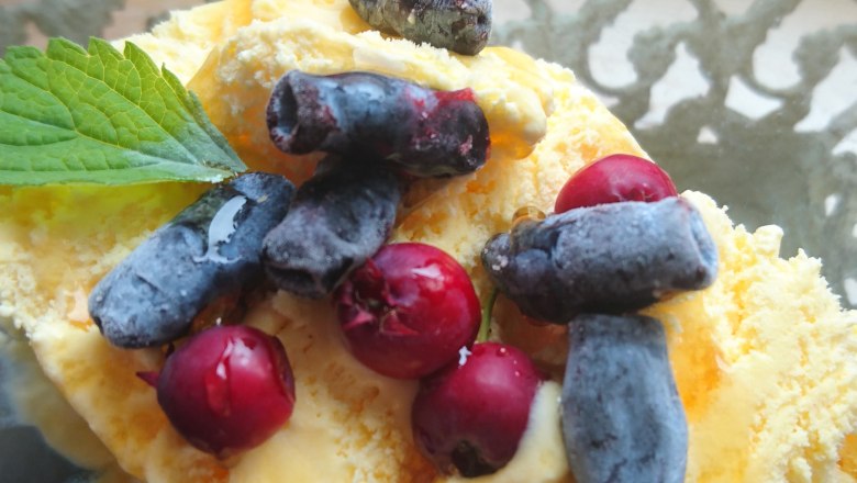 A dessert with yellow ice cream, red and blue berries, garnished with a green leaf, in a glass bowl.