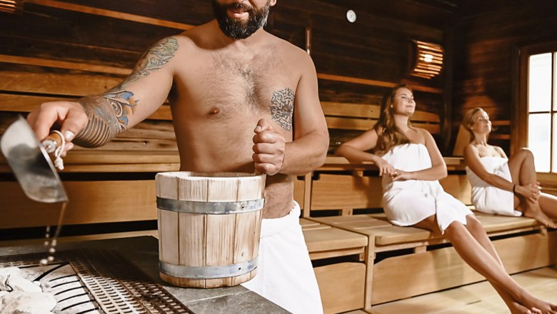 Mann gie&szlig;t Wasser in Sauna, zwei Frauen sitzen entspannt.