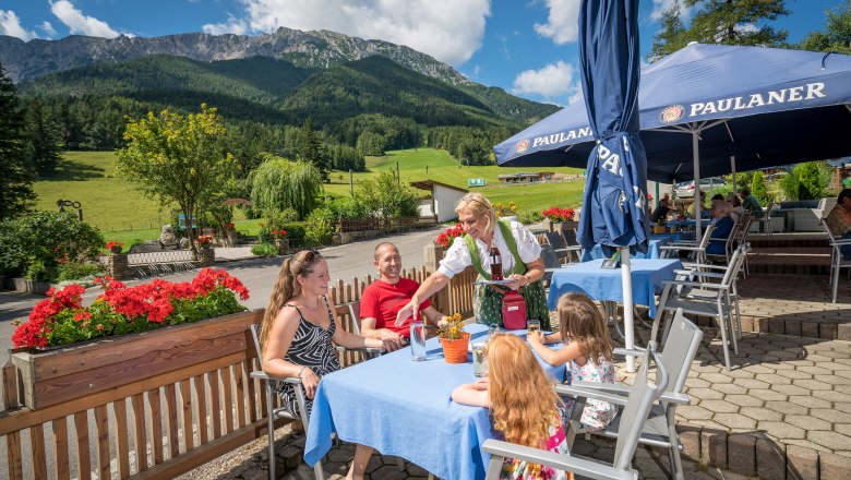 Family on a sunny terrace with mountains in the background, served by a waitress in traditional dress.